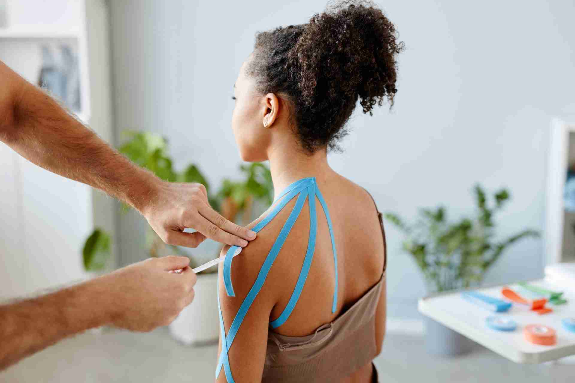Close up image of therapist putting k tape on shoulder of young African American woman in physiotherapy clinic